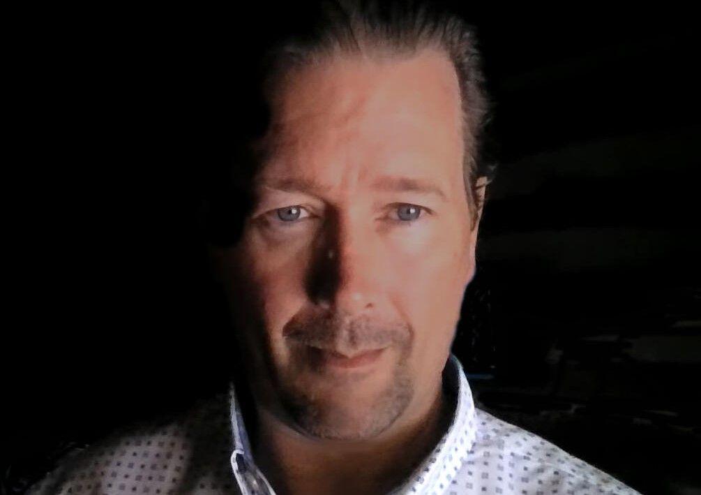 Person wearing a patterned button-up shirt in a dimly lit environment.